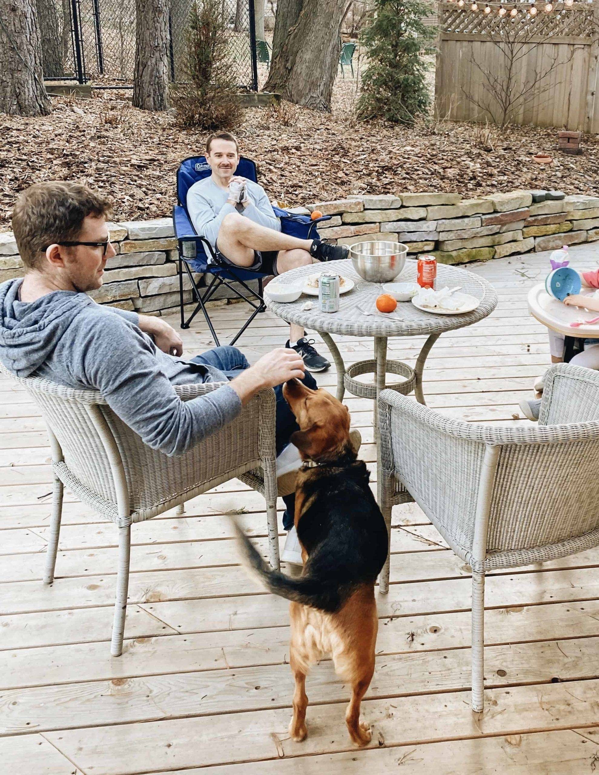 Two men sitting in patio chairs, one man petting a black and brown dog.