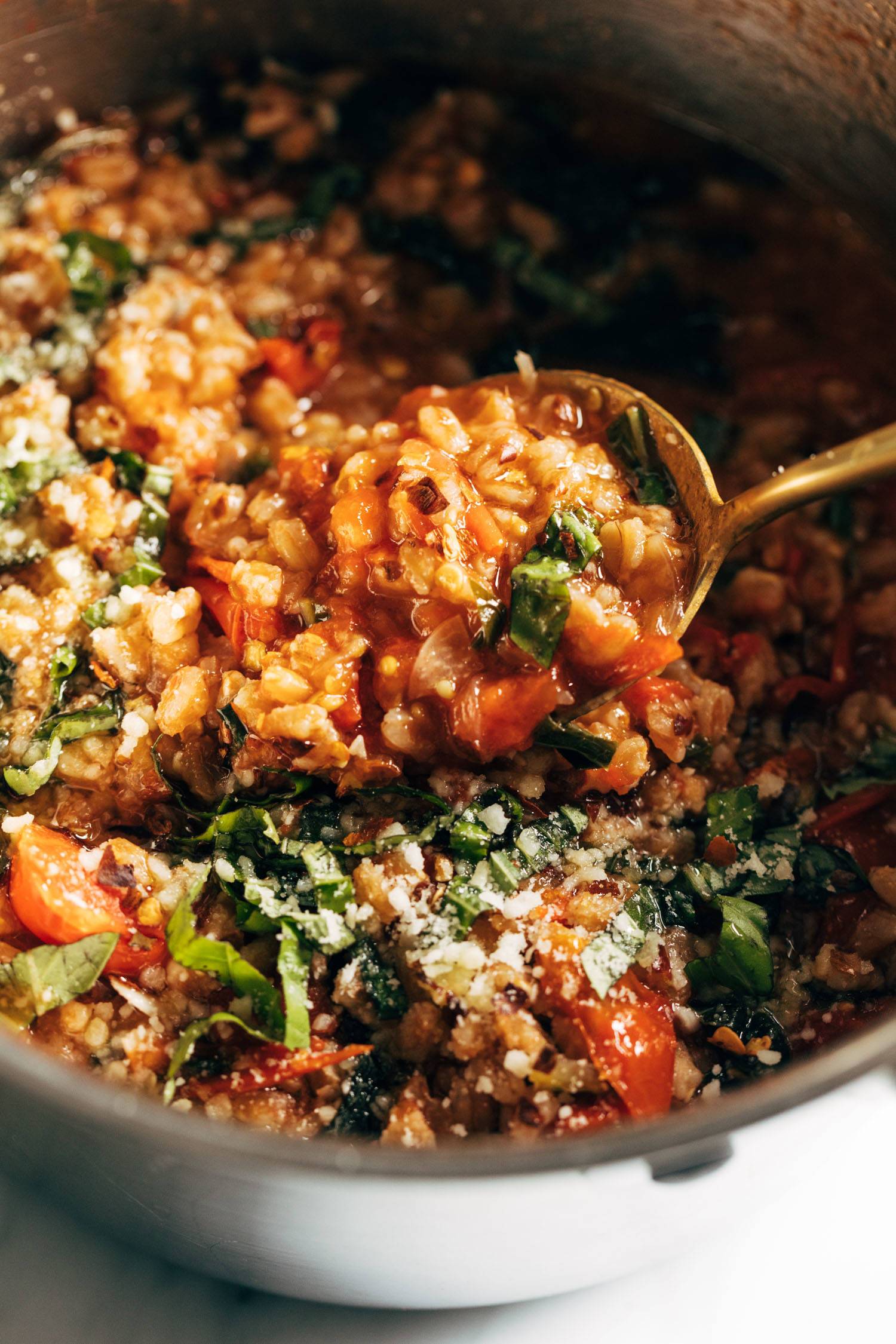 Gold spoon scooping up farro with tomatoes and kale. There are red pepper flakes on top.