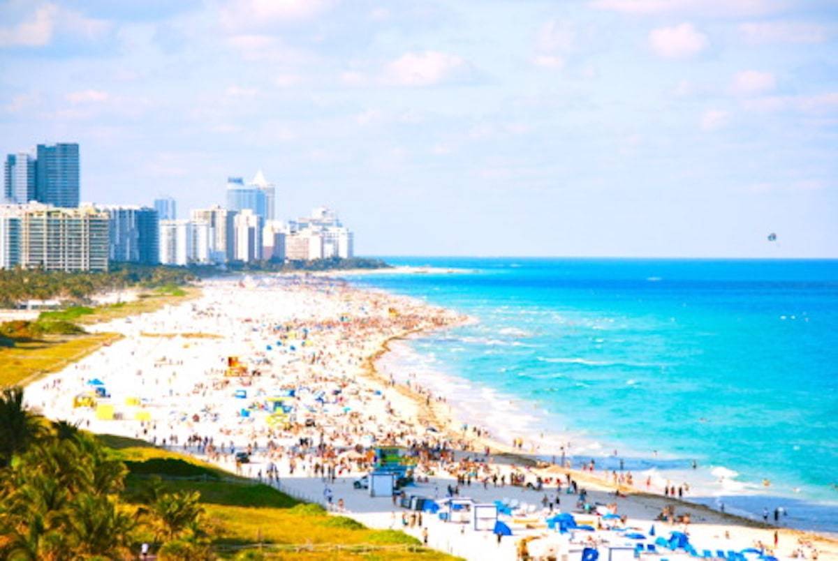 Ocean shore with people filling the sand near various buildings.